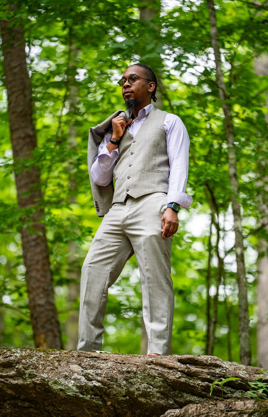 Man in a gray three-piece suit standing on a fallen log in a green forest