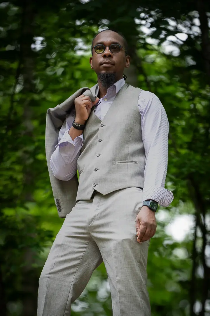 Man in round tinted glasses and a gray three-piece suit facing the camera in a green forest