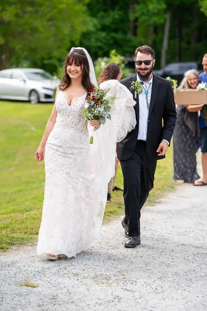 Bride with bouquet and smiling groom walking a gravel path with guests behind