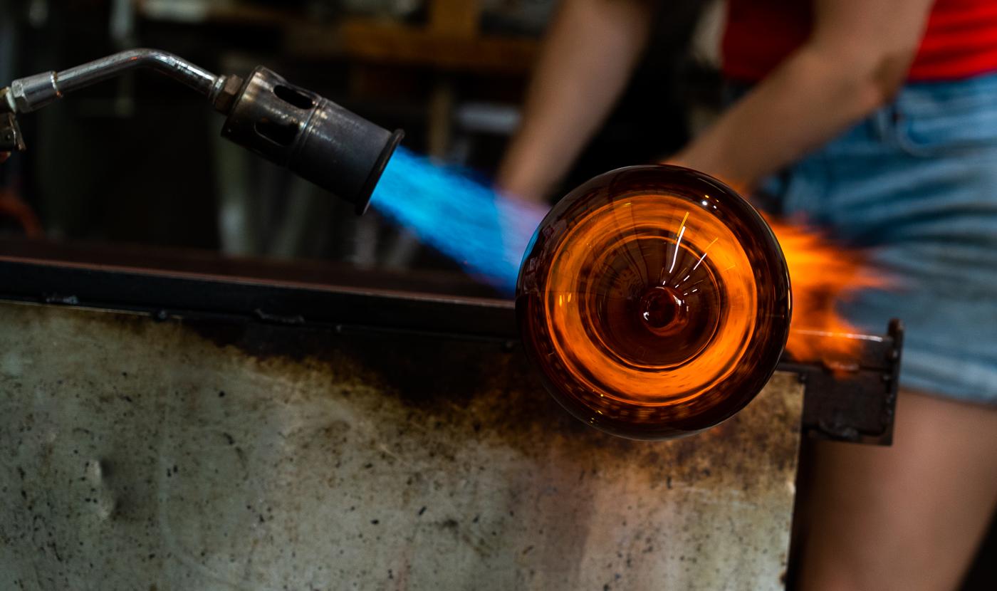 Blowtorch with blue flame heating a glowing amber glass vessel in a glass studio