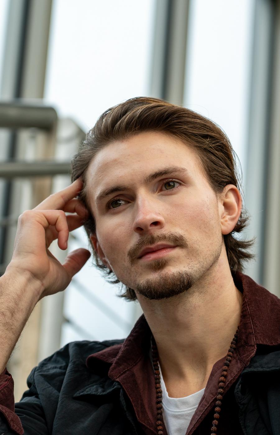 Young man with swept-back hair posing thoughtfully, glass architecture behind him