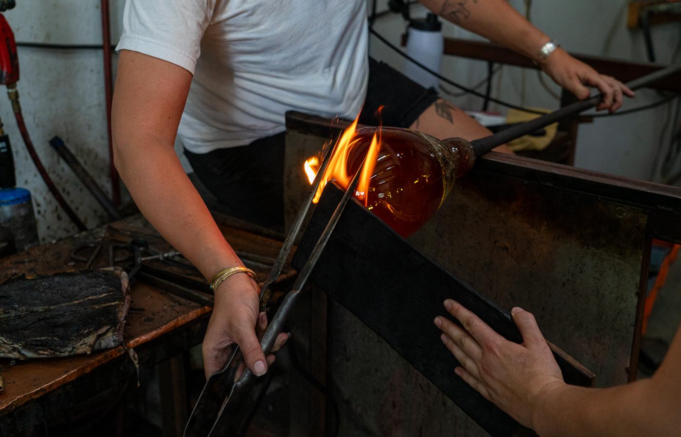 Two glass artisans shaping molten amber glass with flames rising in a workshop